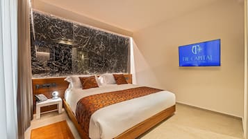 Una habitación de hotel moderna con una cama grande, cabecera de mármol, mesita de noche con teléfono y televisión mostrando el nombre del hotel.
