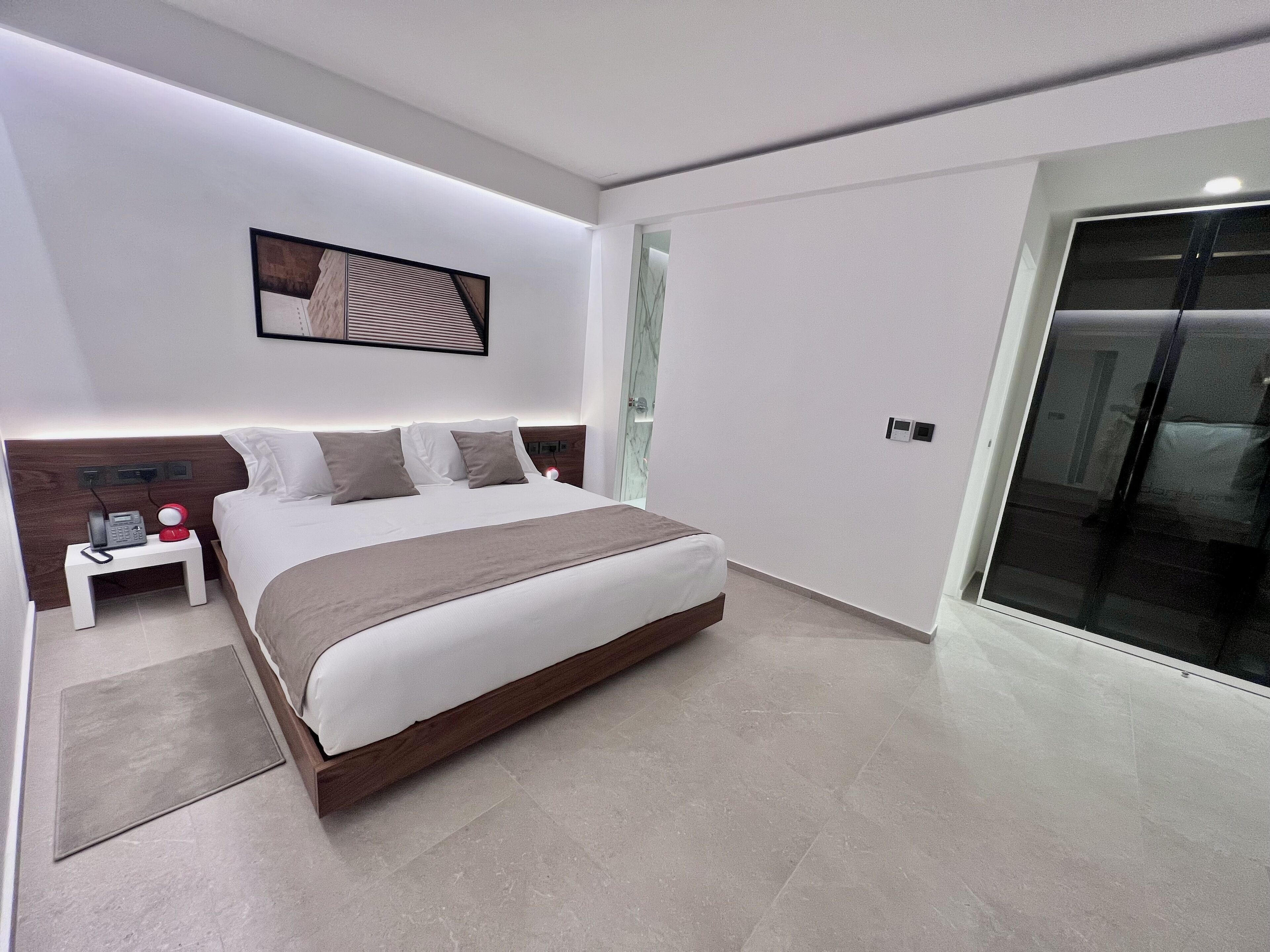 A modern bedroom with a large bed, bedside tables, a nightstand, a wall-mounted phone, and a door leading to another room.