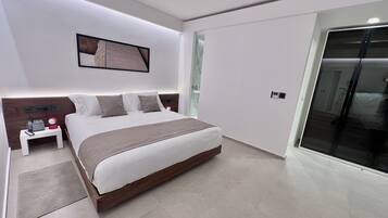 A modern bedroom with a large bed, bedside tables, a nightstand, a wall-mounted phone, and a door leading to another room.