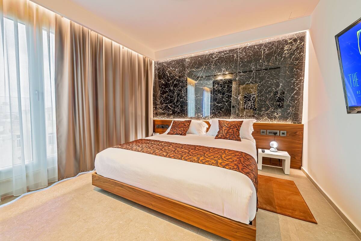 A modern hotel room with a large bed, a marble accent wall, a nightstand with a lamp, and a TV mounted on the wall.