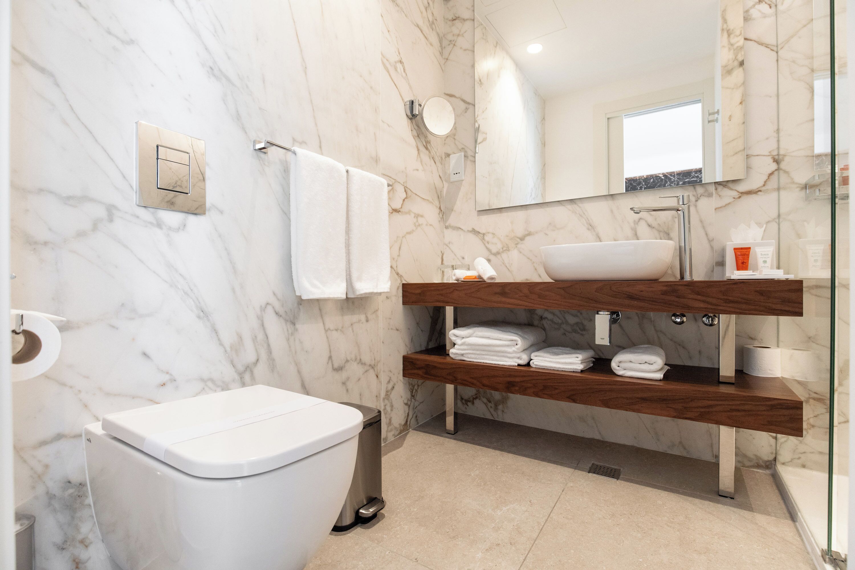 Un baño moderno con pared de mármol, inodoro blanco, tocador de madera con lavamanos y un espejo.