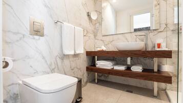 Un baño moderno con pared de mármol, inodoro blanco, tocador de madera con lavamanos y un espejo.