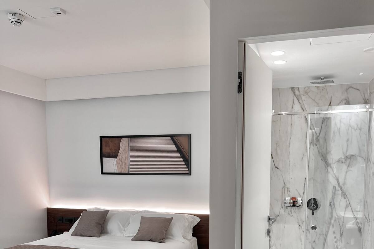 A modern bedroom with a bed, a bedside table, a wall-mounted picture, and a bathroom with a shower and toilet.