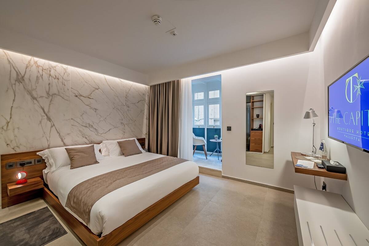 A modern hotel room with a marble wall, a large bed, a desk with a computer, and a TV displaying hotel information.