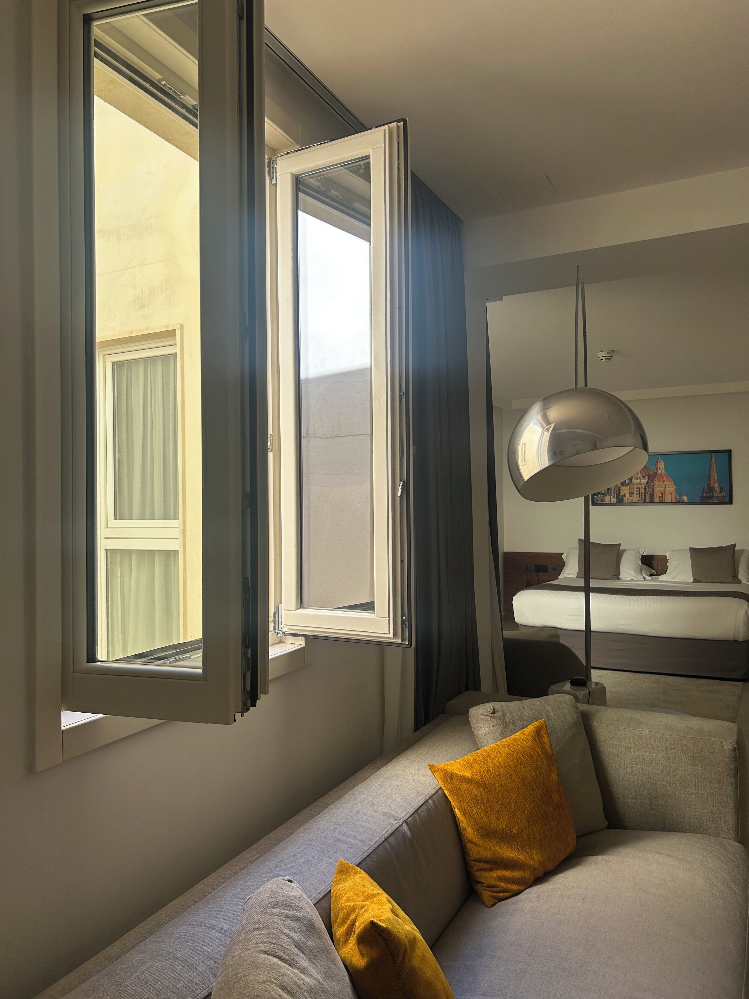 A hotel room with a large window, a sofa with cushions, a bedside table, and a lamp.
