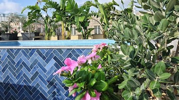 Una piscina con paredes de azulejos azules, rodeada de diversas plantas verdes y flores rosadas.