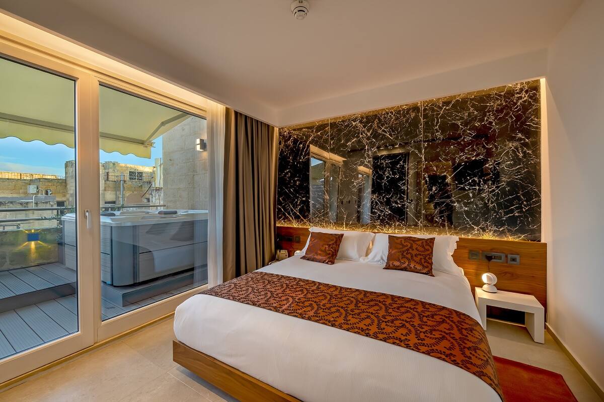 A modern hotel room with a large bed, a glass door leading to a balcony, and a marble wall.