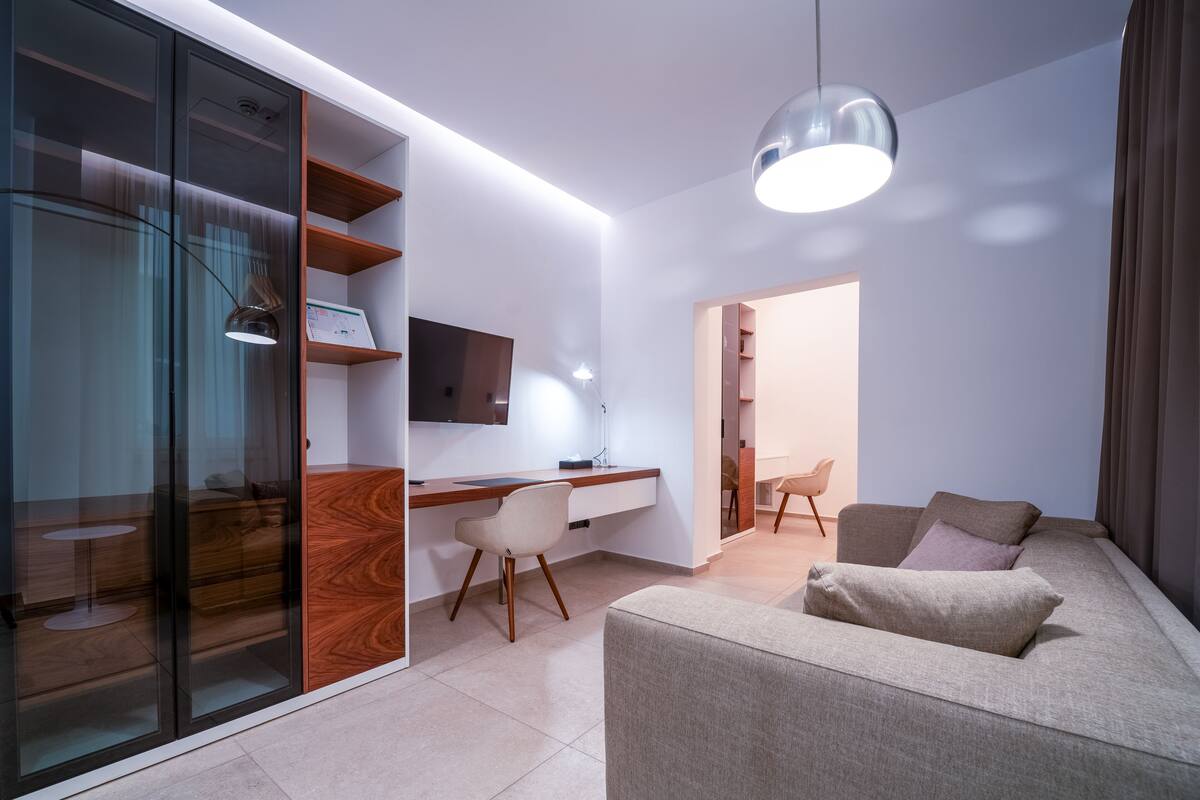 A modern living room with a sofa, a desk, and a TV. There is a glass partition with shelves and a lamp.