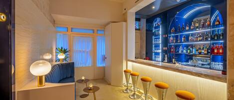 Zona de bar con una estantería curva que exhibe botellas, un área de descanso con sofá y mesas redondas, y vista al exterior a través de amplios ventanales.
