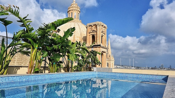 Una piscina con borde de azulejos, un edificio grande con cúpula y campanario al fondo, y un cielo despejado con nubes.