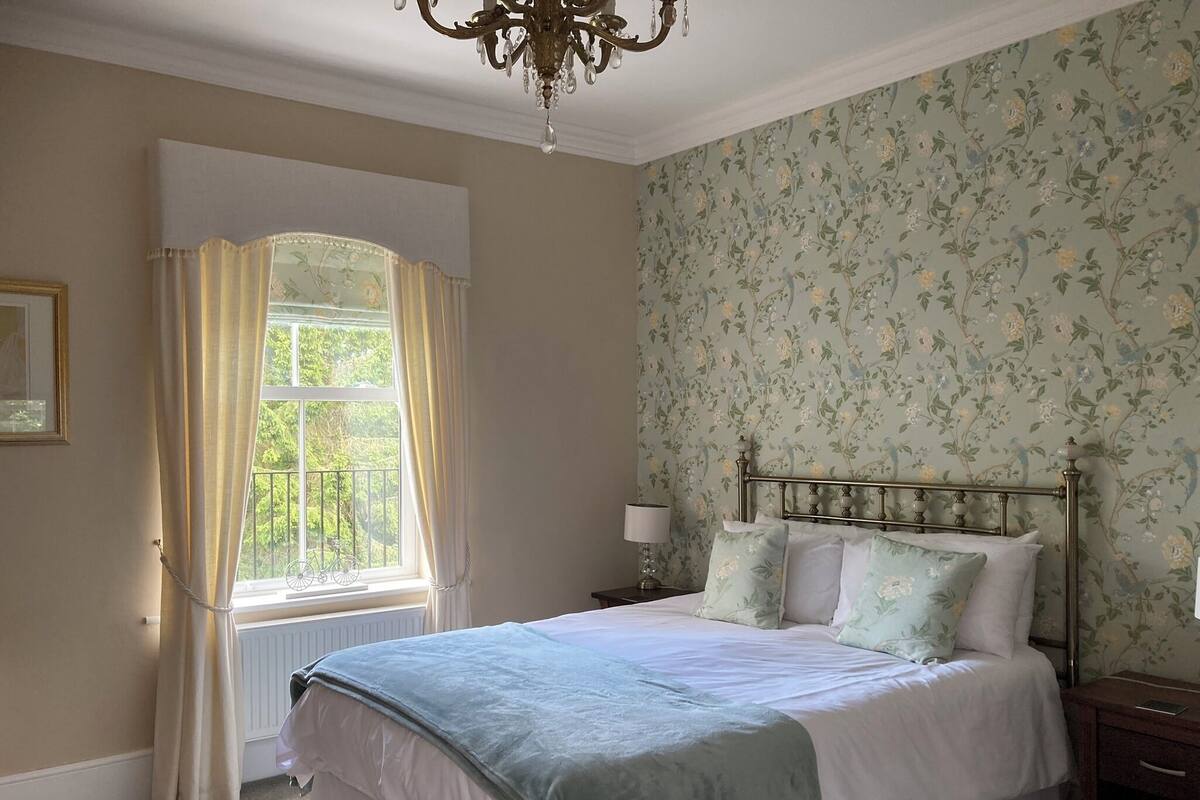 A bedroom with a bed, a nightstand, a lamp, a window with curtains, and a floral wallpaper.