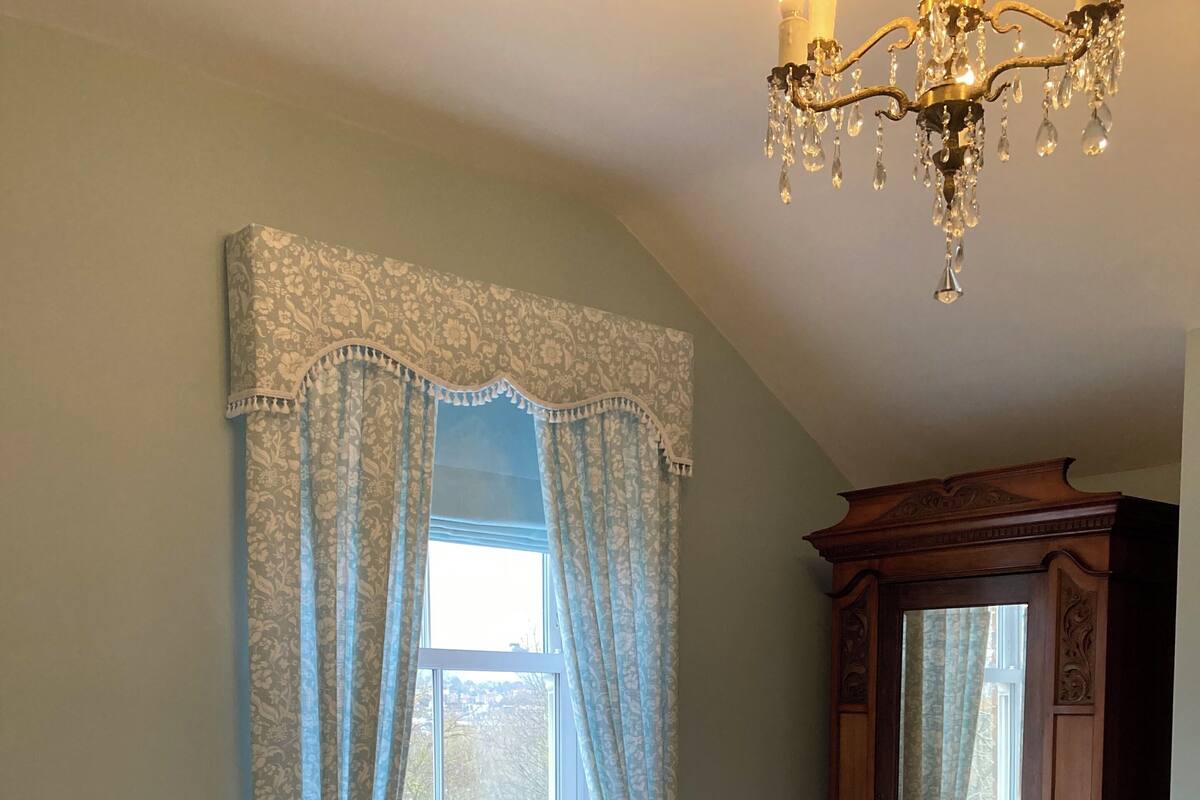 A bedroom with two beds, a large window with curtains, a radiator, a wooden wardrobe, and a chandelier.