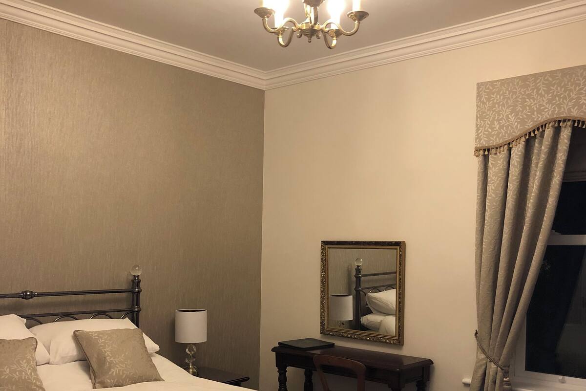 A neatly arranged bedroom with a large bed, a bedside table with a lamp, a mirror, and a window with curtains.