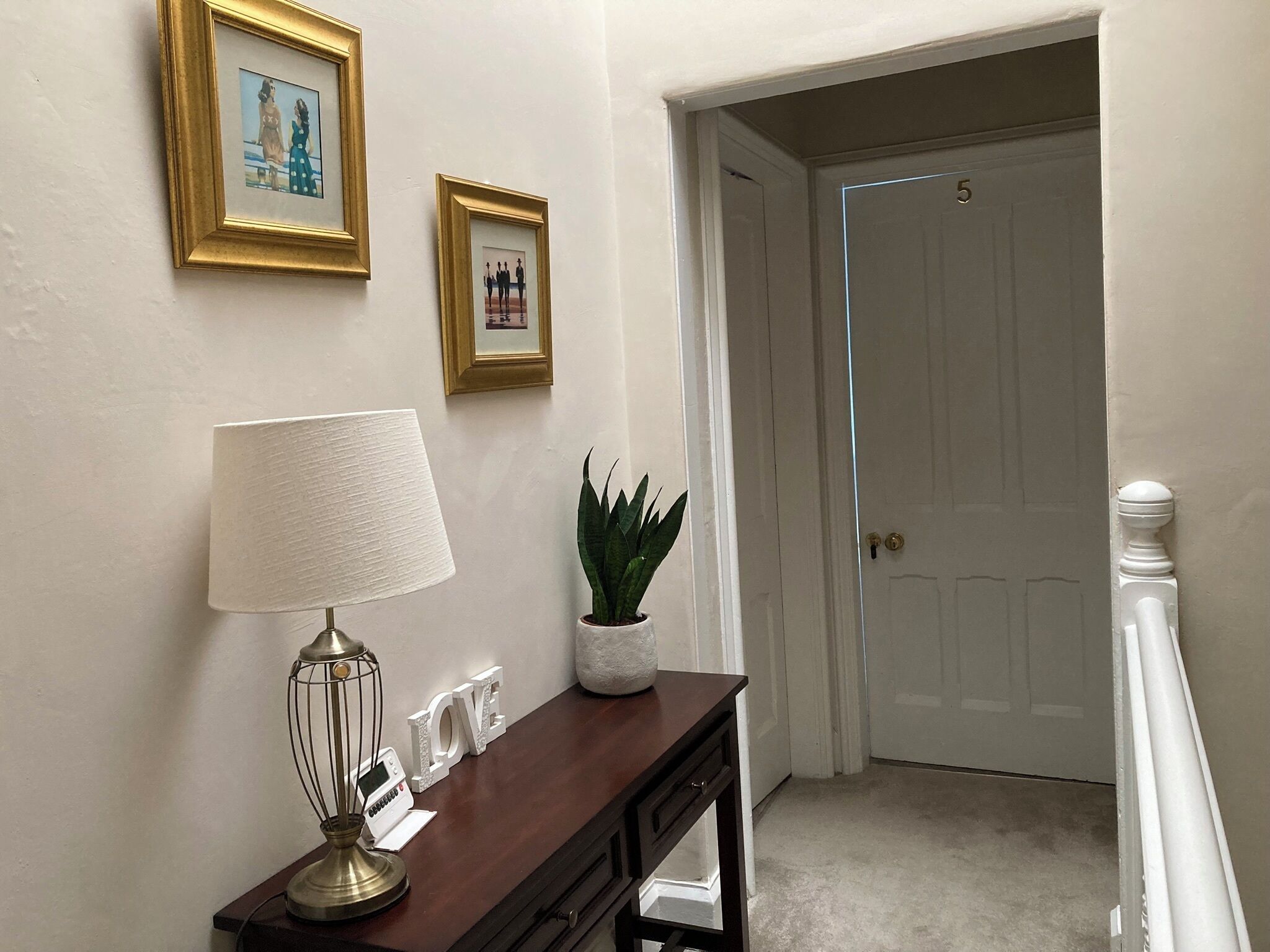 A hallway with a desk, a lamp, framed pictures, and a potted plant.