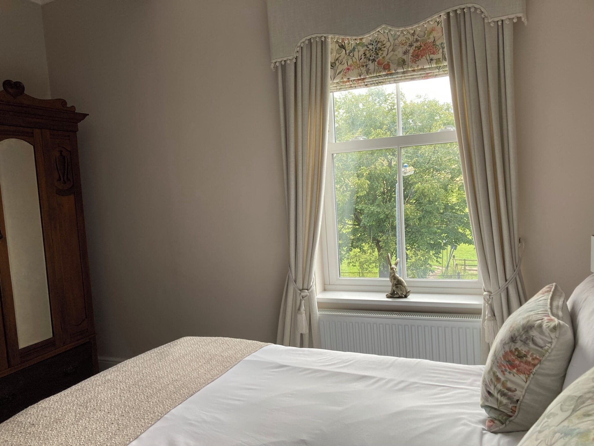 A bedroom with a large window, a bed with white linens, a wooden dresser, and a small statue on the windowsill.