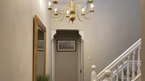 Un pasillo con candelabro, espejo, cuadro enmarco, planta y escalera.