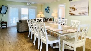A dining area with a wooden table and white chairs, a sofa, a TV, and a painting on the wall.