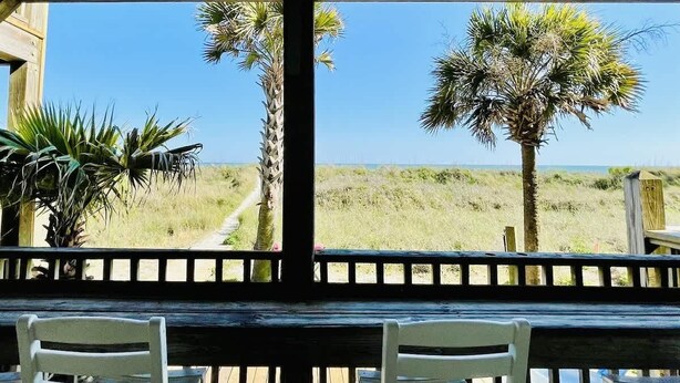 A covered outdoor area with a wooden bench and chairs, palm trees, and a view of the beach.