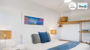 Une chambre d’hôtel comprenant un lit, deux tables de chevet, une petite table, une étagère murale avec des serviettes et un tableau représentant un phare.
