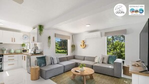 Un salon moderne avec un canapé d’angle gris, une table basse ronde et un coin cuisine doté de meubles blancs et d’une fenêtre donnant sur un espace verdoyant.