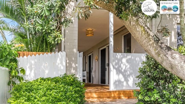 Une maison avec une clôture blanche en bois, un porche en bois et un grand arbre.