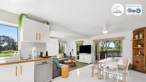 Une cuisine moderne avec des meubles blancs, un lave-vaisselle et une machine à café. On y trouve un coin repas avec une table et des chaises, ainsi qu’une télévision dans le salon.