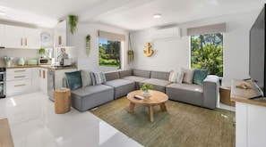Ein modernes Wohnzimmer mit einem grauen Ecksofa, einem runden Couchtisch und einer Küchenzeile mit weißen Schränken und einem Fenster mit Blick auf Grünflächen.