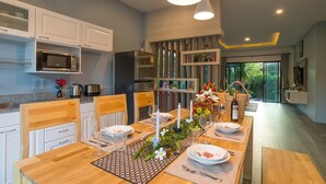 A modern kitchen with white cabinets, a built-in microwave, and a dining area set for six with a wooden table, chairs, and a centerpiece. - Love ao nang pool villa  (อ่าวนาง)