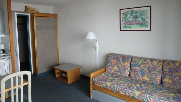 Een kleine kamer met een houten garderobe, een tafeltje, een kleurrijke bank, een lamp en een lijst met een afbeelding aan de muur.