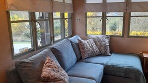 A room with a sofa, patterned pillows, a lamp, and large windows with a view of trees and bushes. - The Meuse is at the end of the garden (Fépin)
