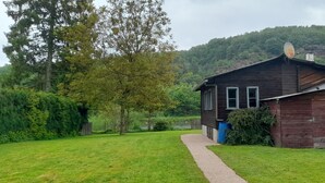 A rustic wooden cabin with a gravel path leading to it, surrounded by lush greenery and a forested hillside. - The Meuse is at the end of the garden (Fépin)