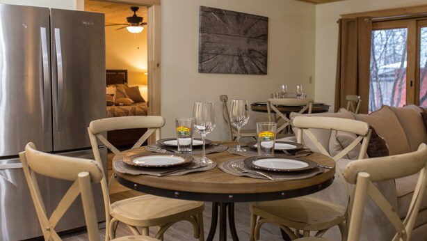 A dining area with a round table set for four, surrounded by four chairs. A large window with curtains is visible in the background.