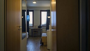 Et hotelværelse med trægulv, en seng, et skrivebord med stol, et lille køleskab og et vindue med gardiner.