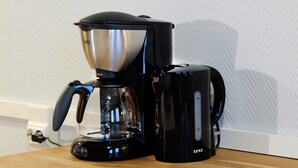En kaffemaskine i sort farve med en gennemsigtig vandbeholder og en sort drypbakke, placeret på en træoverflade.