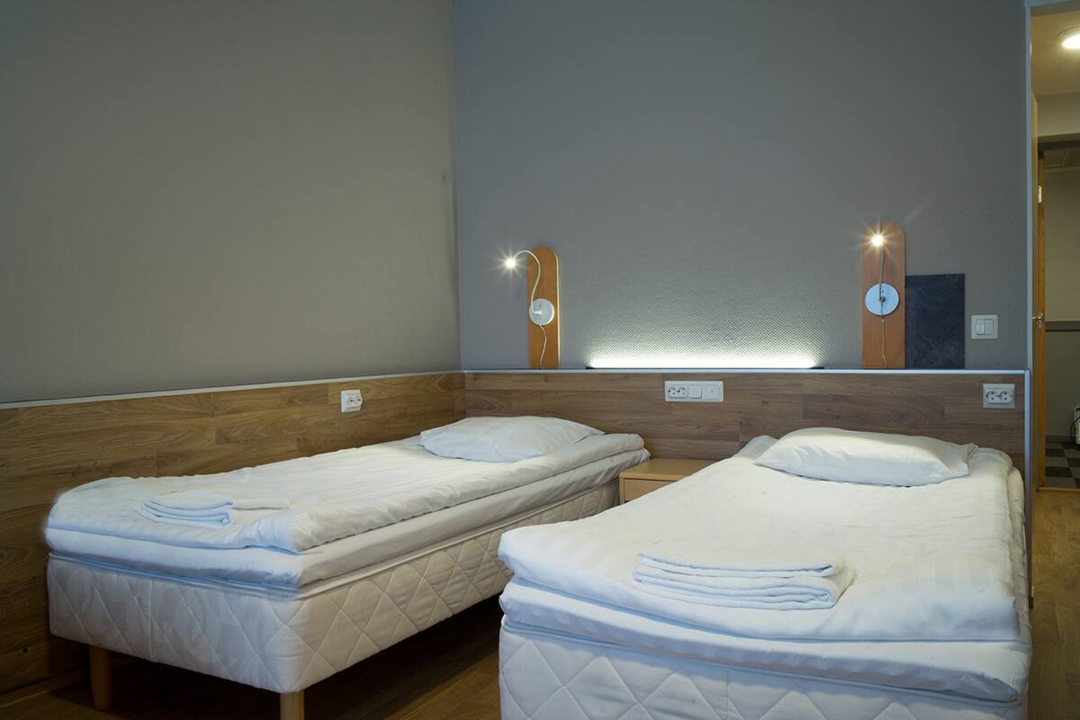 Deux lits simples avec des têtes de lit en bois, un miroir et des lampes fixées au mur dans une chambre d’hôtel.