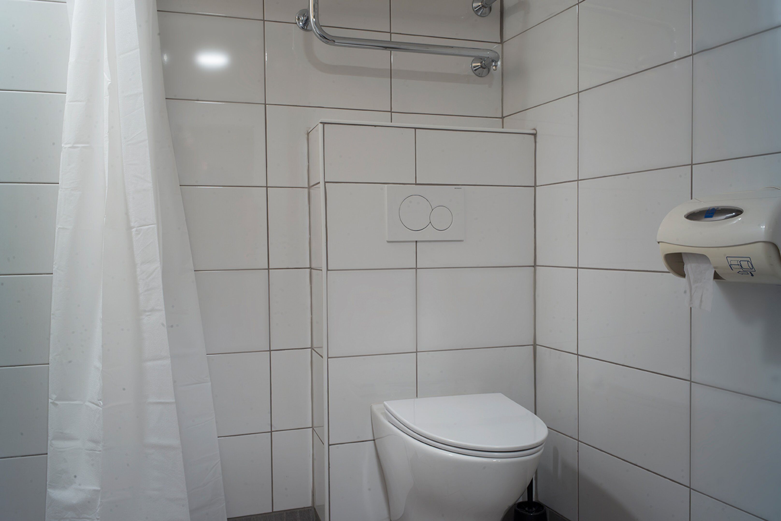 A bathroom with white tiles, a white toilet, a shower area, and a hand sanitizer dispenser.