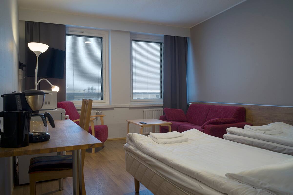 Une chambre d’hôtel avec un lit, un bureau doté d’une machine à café, une chaise, un canapé rouge et un téléviseur fixé au mur.