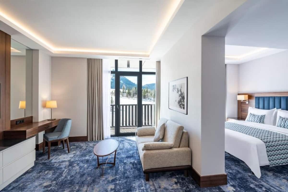 A hotel room with a bed, a sofa, a desk, a chair, and a view of the outdoors.