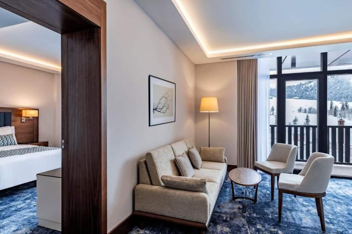 A hotel room with a bed, a sofa, two armchairs, a small table, and a large window with a view of a snowy landscape.