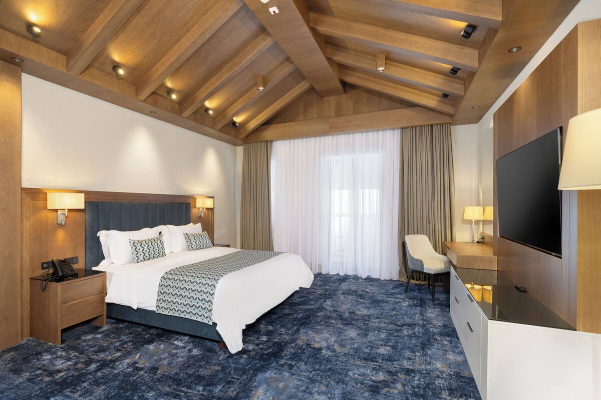 A hotel room with a bed, bedside table, lamp, TV, and a large window with curtains.