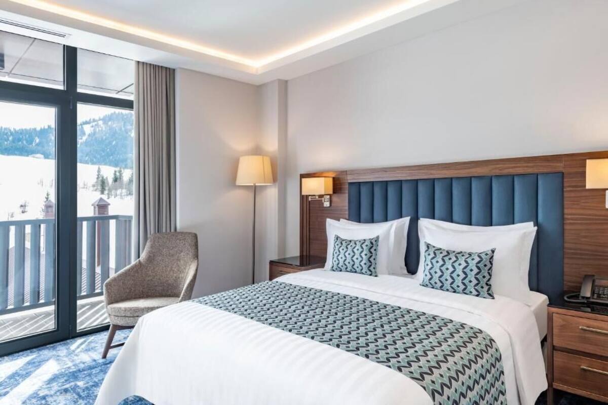 A hotel room with a large bed, a chair, a nightstand, and a view of the outdoors.