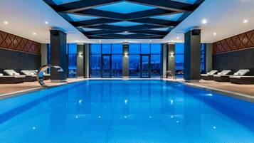 Una amplia piscina interior con un diseño geométrico en el techo, grandes ventanales y tumbonas a lo largo de los bordes.