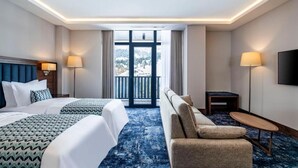 Ein Hotelzimmer mit zwei Betten, einer Sitzecke, einem kleinen Tisch und einem Fernseher. Es gibt einen Balkon mit Blick auf Bäume und Berge.