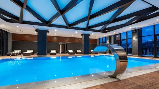 Ein Hallenbad mit klarem, blauem Wasser, Holzboden und modernem Design mit sichtbaren Holzbalken und großen Fenstern.