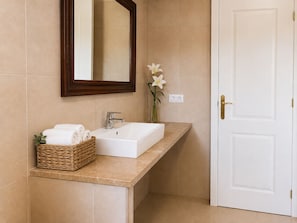 Een badkamer met een witte wastafel, een spiegel, een vaas met een bloem en een mand vol handdoeken.