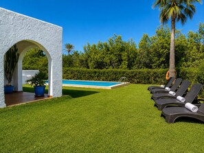 A pool area with a white structure, a palm tree, and a row of lounge chairs.