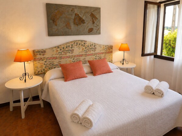 A hotel room with a bed, two bedside lamps, a window with curtains, and a painting on the wall.
