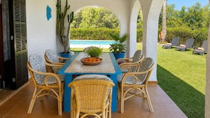 Une terrasse couverte avec une table et des chaises, une piscine en arrière-plan et une pelouse bien entretenue.