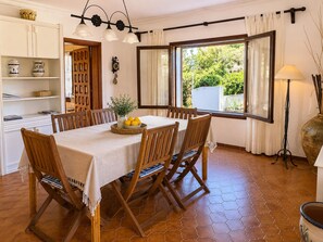 A dining area with a table set for six, surrounded by wooden chairs, a window with curtains, and a view of greenery outside.