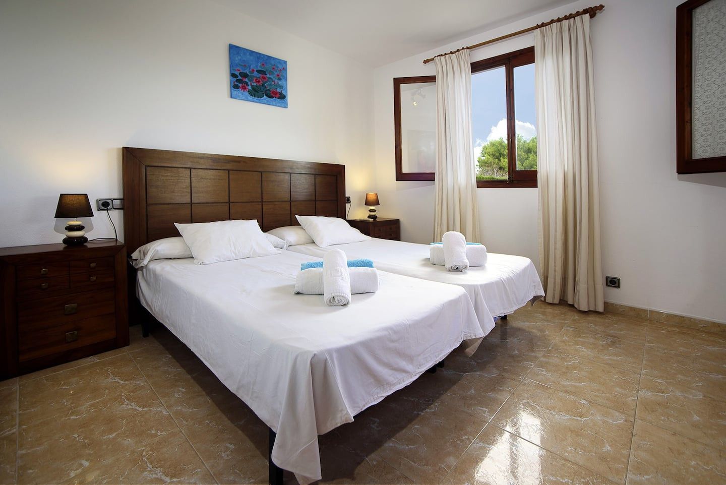 A hotel room with a double bed, two bedside tables, a nightstand, a window with curtains, and a painting on the wall.